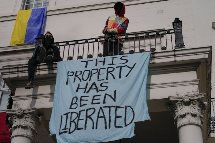 A man sits on the balcony of stately home holding a banner reading This Property Has Been Liberated