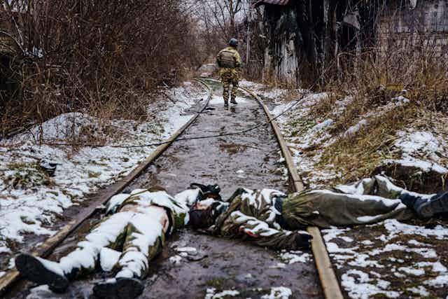 Two dead Russian soldiers, wearing uniforms and covered with snow, lie on railroad tracks as a Ukrainian soldier walks away from them along the track..