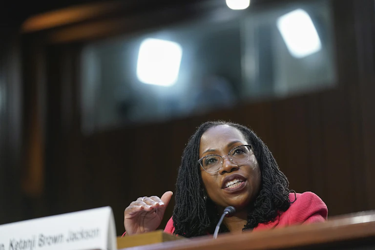A woman sits at a table in front of microphone and gestures as she speaks.