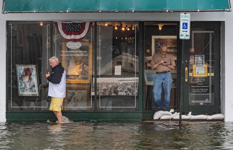 A man stands just inside the glass door of an art gallery with sandbags keeping out ankle-deep water while a shopper wades past.