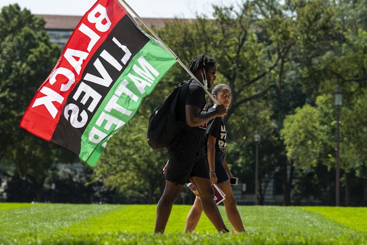 Photo showing two Black activists, one carrying a black backpack and a large 'Black Lives Matter' flag, both people walking on a public lawn in Washington, D.C. with trees in the background. The event was held on the 58th anniversary of the March On Washington.