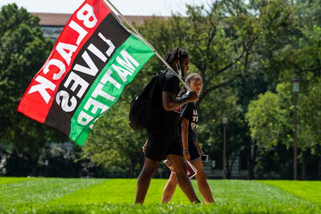 Two young black activists are walking on a grass lawn while one holds a Black Lives Matter flag.