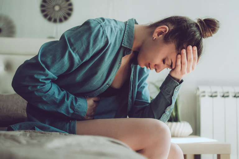 A young woman sitting on her bed bent over, holding her stomach in pain.