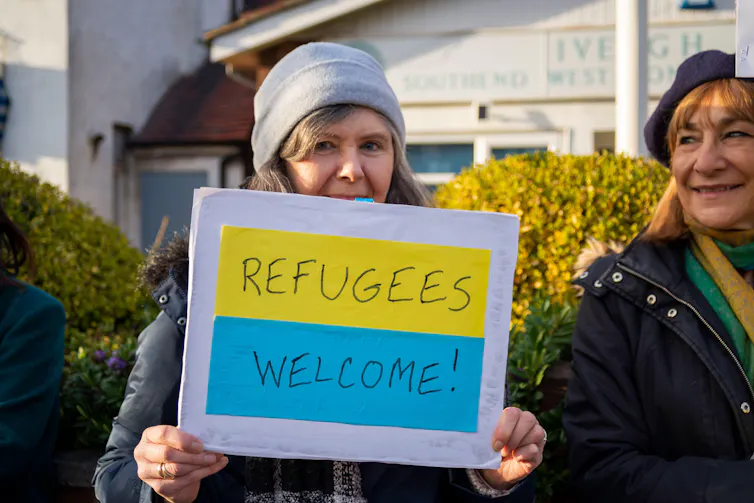A woman holding a sign coloured like the Ukrainian flag reading 'refugees welcome'.
