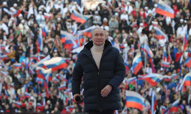 A man in an overcoat and a turtleneck sweater stands in front of a crowd waving Russian flags