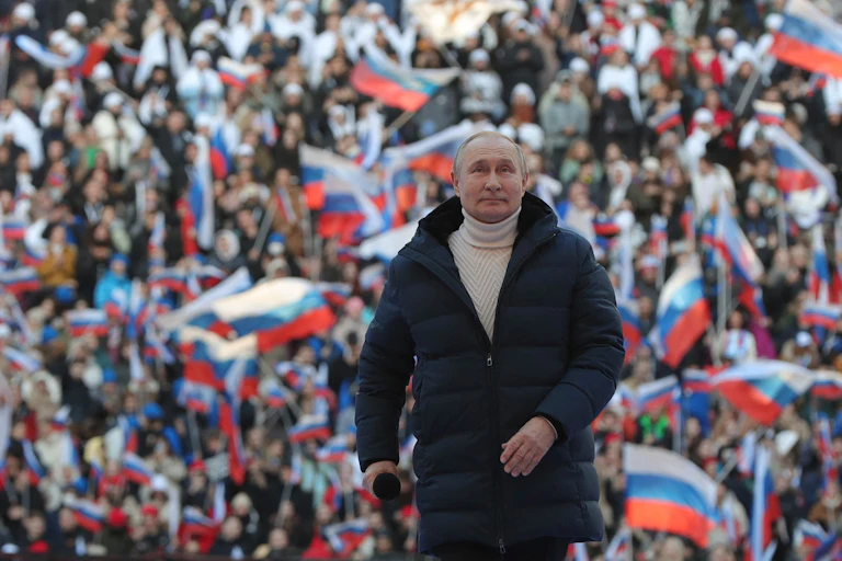 A man in an overcoat and a turtleneck sweater stands in front of a crowd waving Russian flags
