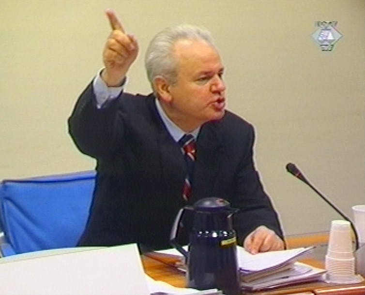 A man in a suit and tie sits at a desk waving his hand in the air