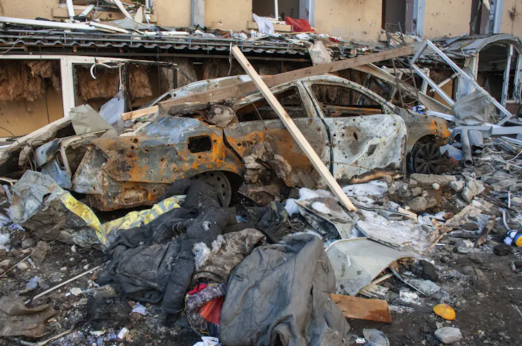 Scène de destruction dans une rue, avec une voiture incendiée en avant-plan