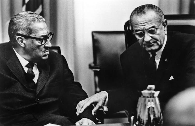 Two men in business suits, one Black and one white, are pictured sitting together in a black and white photo