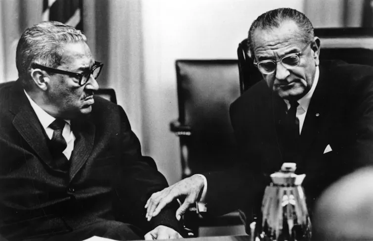 Two men in business suits, one Black and one white, are pictured sitting together in a black and white photo