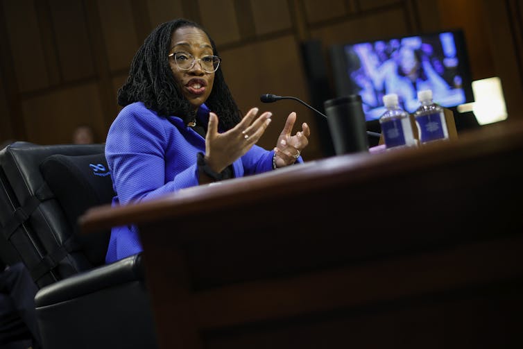 A black woman in a bright blue jacket gestures while speaking into a microphone and seated at a large wooden desk