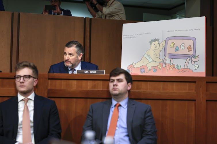 A man at a desk with a plaque reading Mr. Cruz is seated next to a large poster from a kids' book that which shows a white baby playing with blocks that spell out the word race