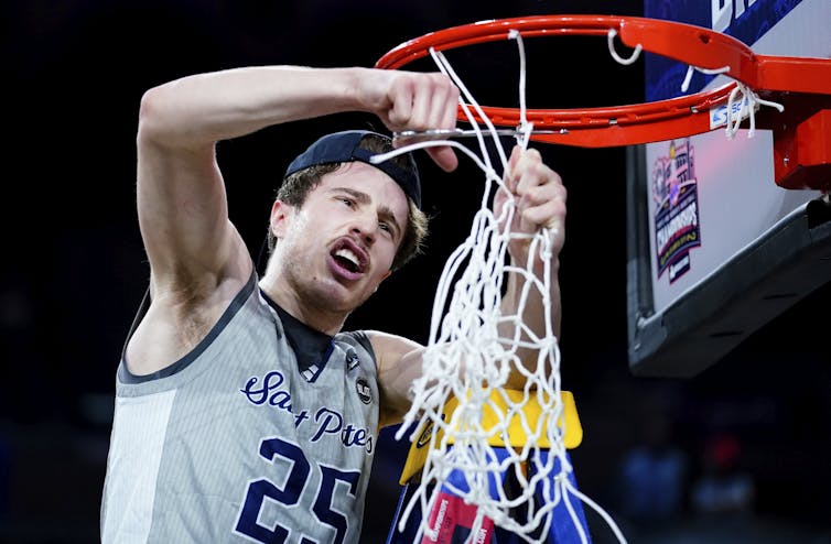 A young white man wearing a saint peter's basketball jersey uses a scissors to cut the net off of a basketball hoop
