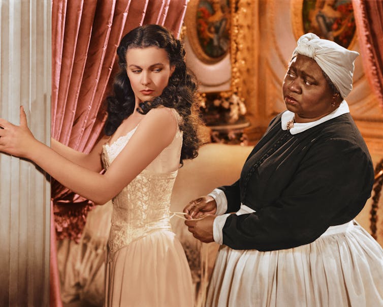 A black woman dressed with a scarf on her head and a white apron is tying the laces of an undergarment worn by a white woman.