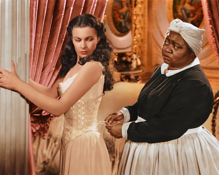 A Black woman dressed with a scarf on her head and a white apron is tying the laces of an undergarment worn by a white woman.