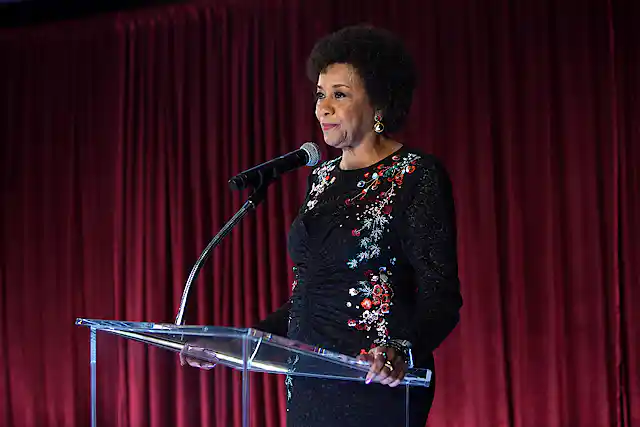 A black woman stands on a stage behind a glass podium and microphone.