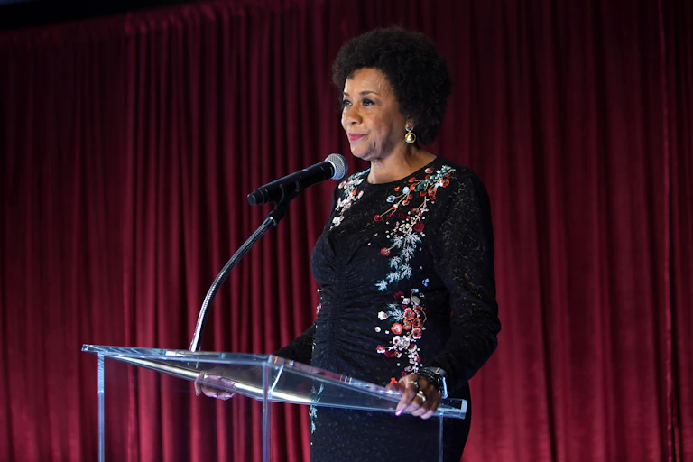 A black woman stands on a stage behind a glass podium and microphone.