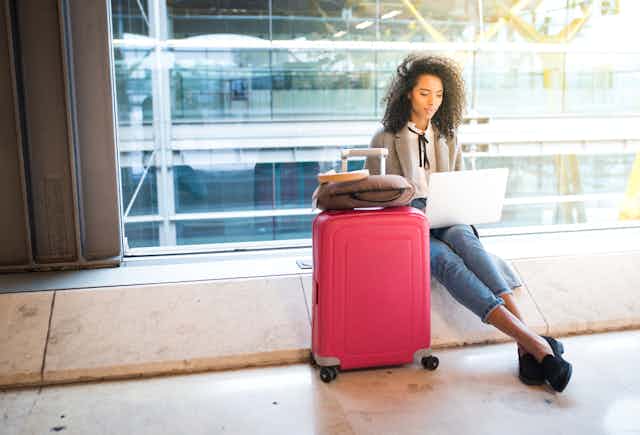 femme noire assise dans un aéroport avec son ordinateur portable et sa valise rouge
