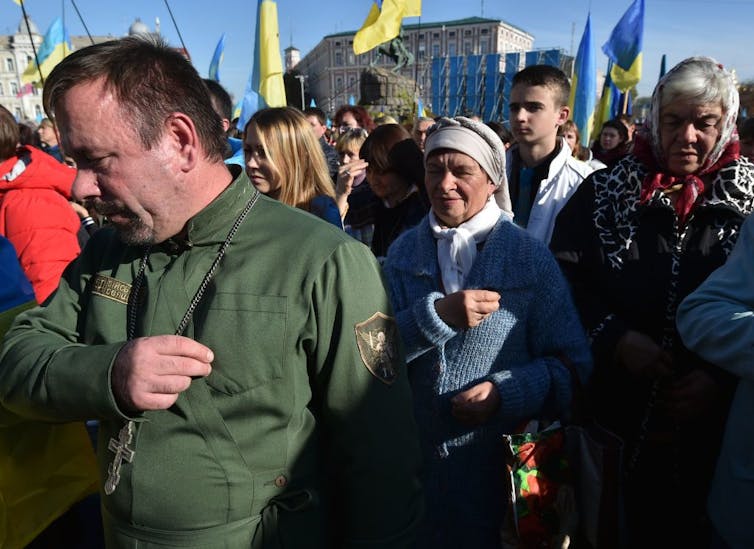 People pray in a large crowd, with Ukrainian flags waving in the background.