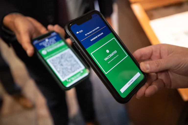 A smartphone displaying the message 'Vaccinated' on a green screen, with another smartphone displaying a QR code in the background