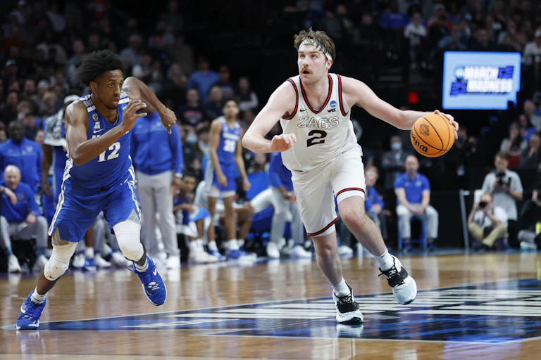 A white man with a mustache holds basketball in his left hand as he dribbles toward the basket and a black man runs toward him on a basketball court
