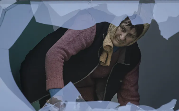 A woman in a kerchief cleans up broken glass surrounded by the shards of a broken window.