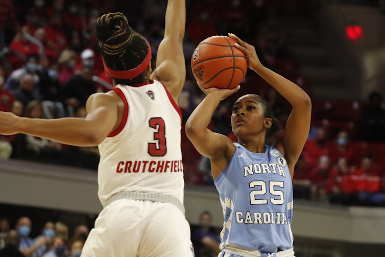 A Black woman wearing a blue jersey jumps as she holds a basketball in her hands and prepares to shoot it over another woman's outstretched arm