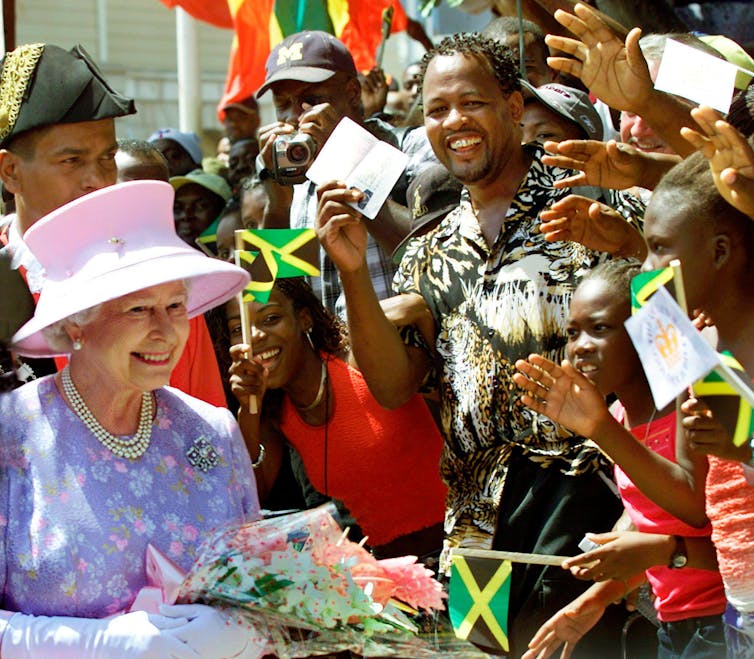 Queen Elizabeth wearing a pink hat smiles and greets a crowd of Jamaican people waving Jamaican flags