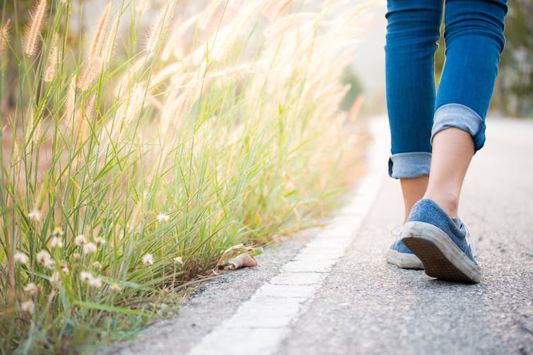 A woman walks past tall grass at the side of a paved path.