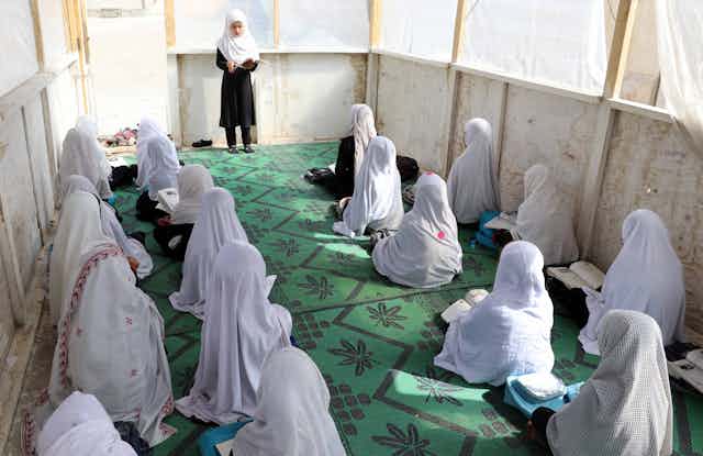 A group of female Afghan primary school pupils sits on the floor of a classroom listening to their female teacher.