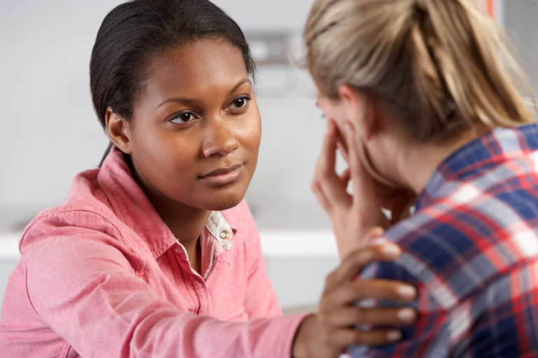 A woman in a pink shirt looks at a woman in a checkered shirt who is crying.