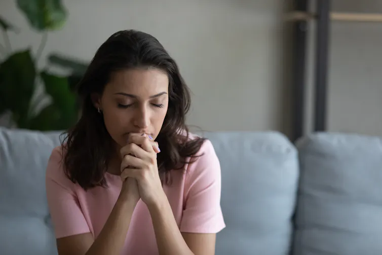 A women holds her hands to her face, with her eyes closed, sitting on a grey couch.