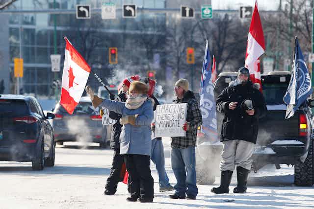 A woman waves as Canada flags hang in the background, a man carries a sign that reads 'imagine there's no mandate, it isn't hard to do