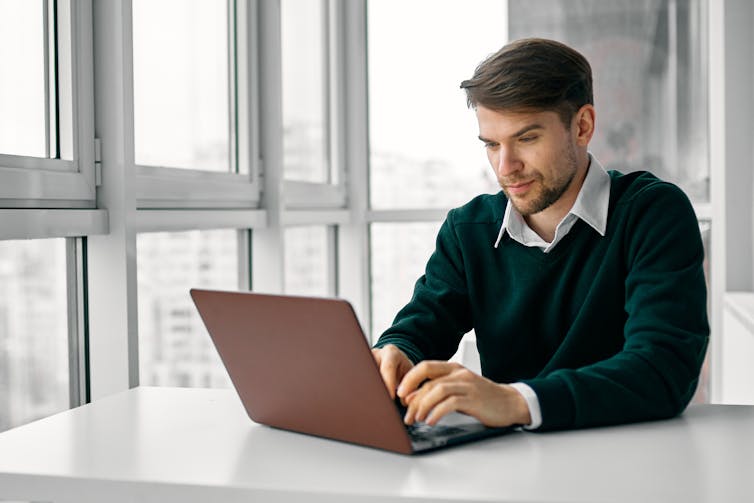 A man wearing a green sweater sits at a white table typing on his laptop.