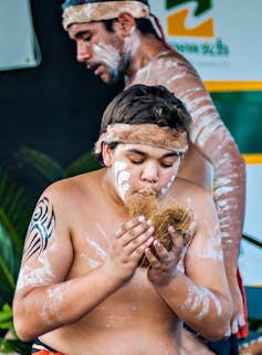 A young First Nations person stands in cultural paint, trying to ignite fire.