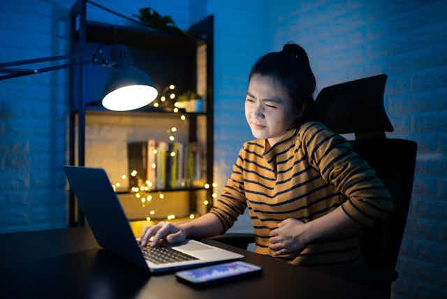 Woman sitting in dark at laptop, clutching her tummy in pain