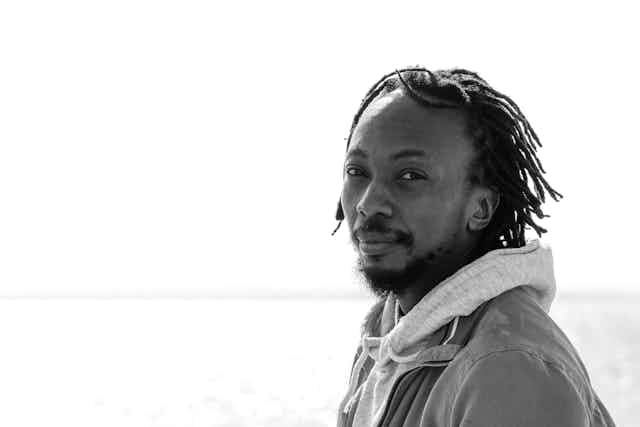 Blak and white portrait of a man with dreadlocks wearing a hoodie with a jacket over; he looks wryly into camera, behind him a distant horizon.
