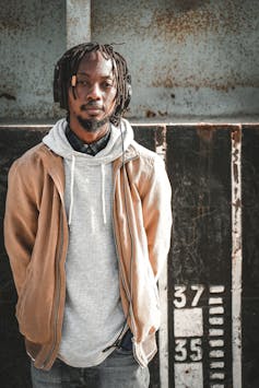 A man stand with his hands in his jacket pockets, in a grey hoodie with dreadlocks; he looks deadpan into camera.