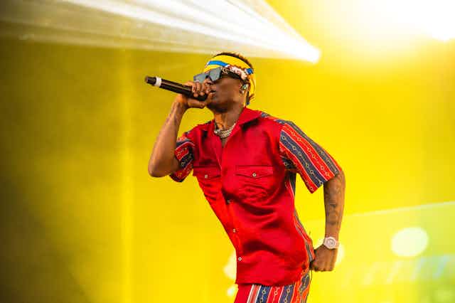 Against a bright yellow backdrop of light, a young man performs into a microphone that he holds up in one hand. He wears bright orange clothing, a bandana in his hair and has sunglasses on.