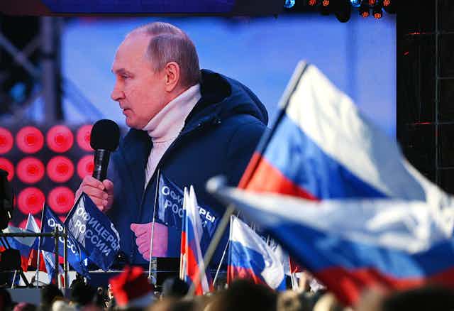 Vliadmir Putin appears on a video screen, making a speech at a rally with a Russian flag in the foreground.
