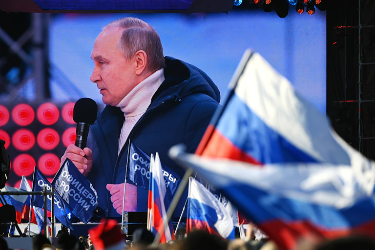 Vliadmir Putin appears on a video screen, making a speech at a rally with a Russian flag in the foreground.