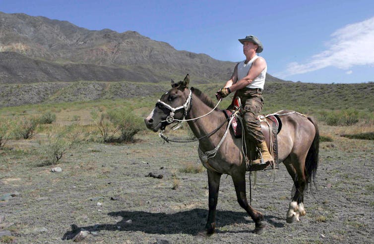 Vladimir Putin on cowboy hat and boots, sits astride a horse.