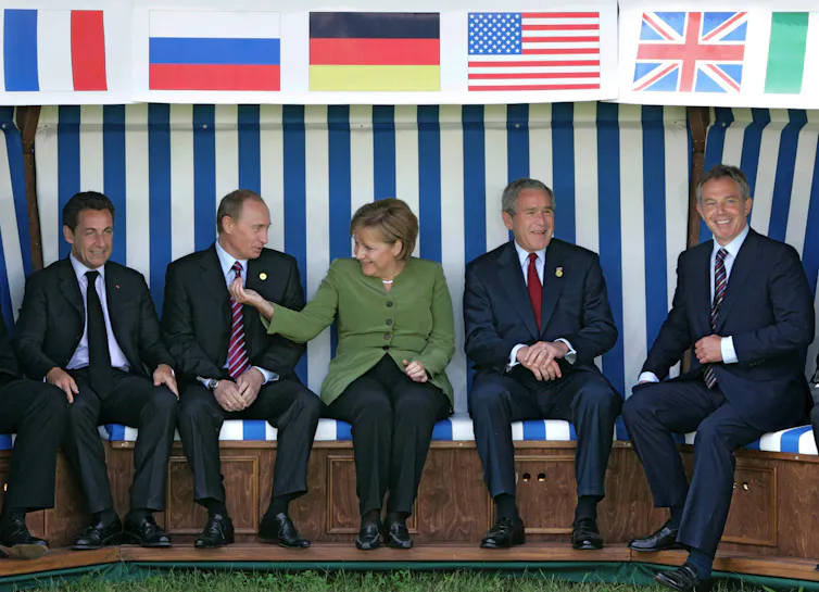 Nicolas Sarkozy, Vladimir Putin, Angela Merkel, George W Bush and Tony Blair sit smiling on a bench under their countries' flags.