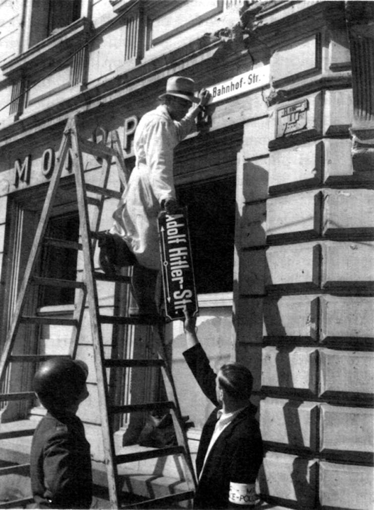 A black and white photograph of a man on a ladder replacing a street sign on the side of a building.