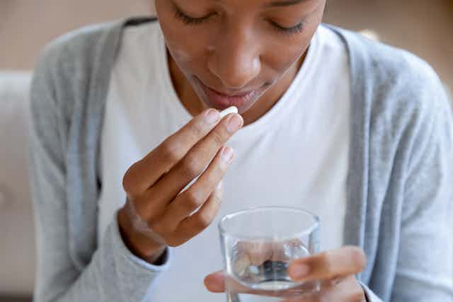 Woman holds a pill up to her mouth in order to take it. She holds a glass of water in her other hand.