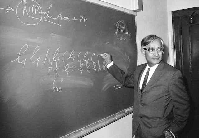 Black and white photo of suited man standing at a blackboard, chalk in hand, writing what looks like genetic sequences.