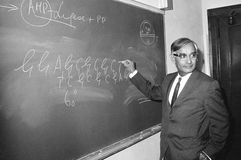 Black and white photo of suited man standing at a blackboard, chalk in hand, writing what looks like genetic sequences.