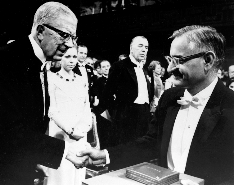 Black and white photo of two men in tuxedos shaking hands. Khorana, on the right, holds what looks like a box possibly containing a medal.