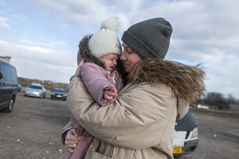 A woman in a beige coat and grey hat colds a young crying baby, in a pink snow suit and grey hat.