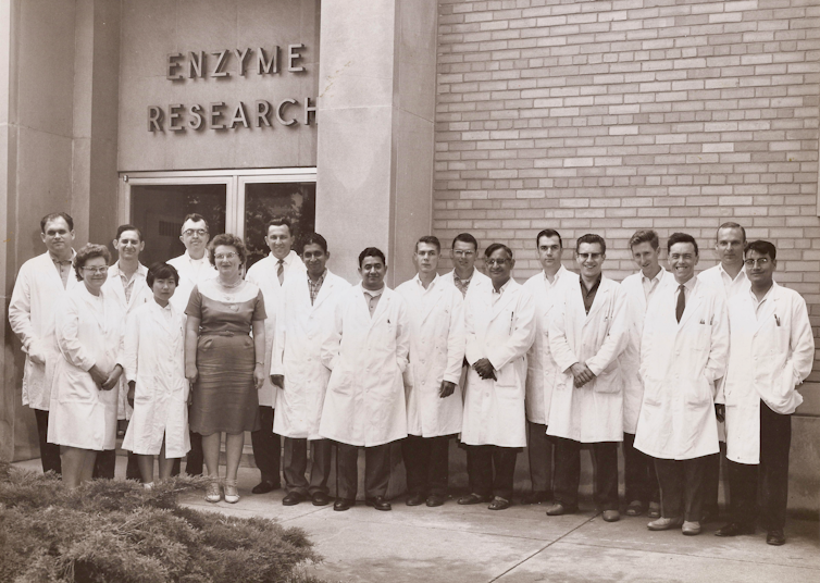 Black and white photo of 18 people, all but one in white lab coats, lined up outside a building labeled 'Enzyme Research.'